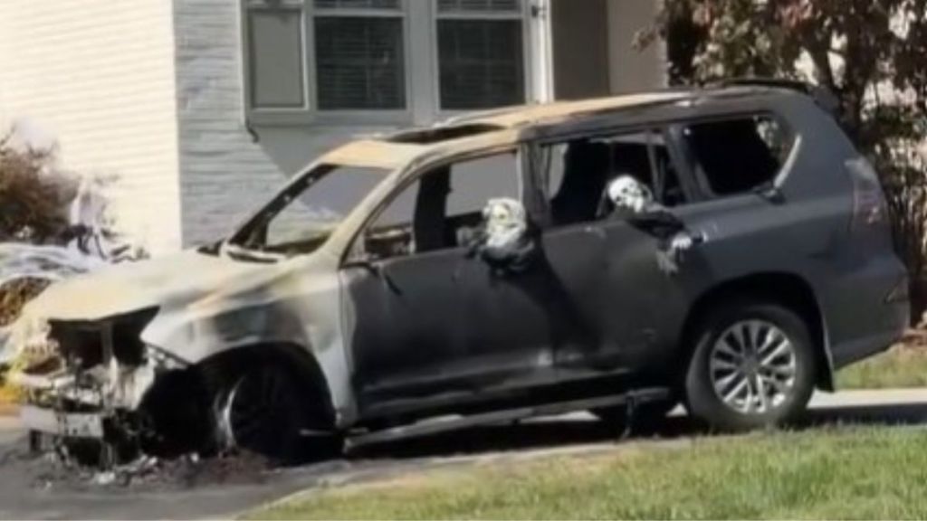 Image shows a burned car sitting in a driveway with Halloween decoration skeletons placed in the passenger compartment.