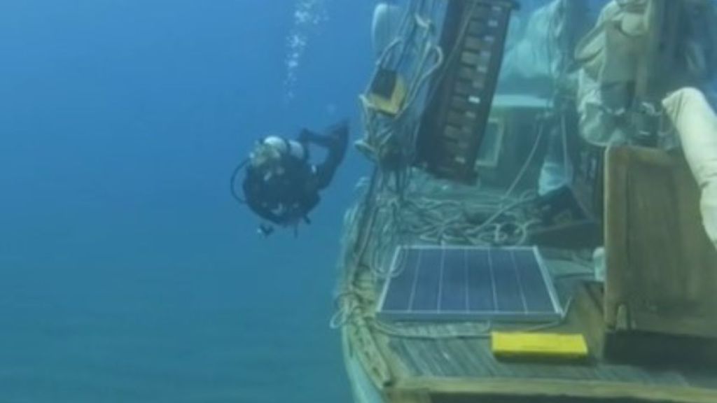 Image shows a diver swimming underwater near a sunken sailboat.