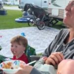 Image shows a family at a campground with a mom rocking holding one screaming child while a toddler has a tantrum next to her.