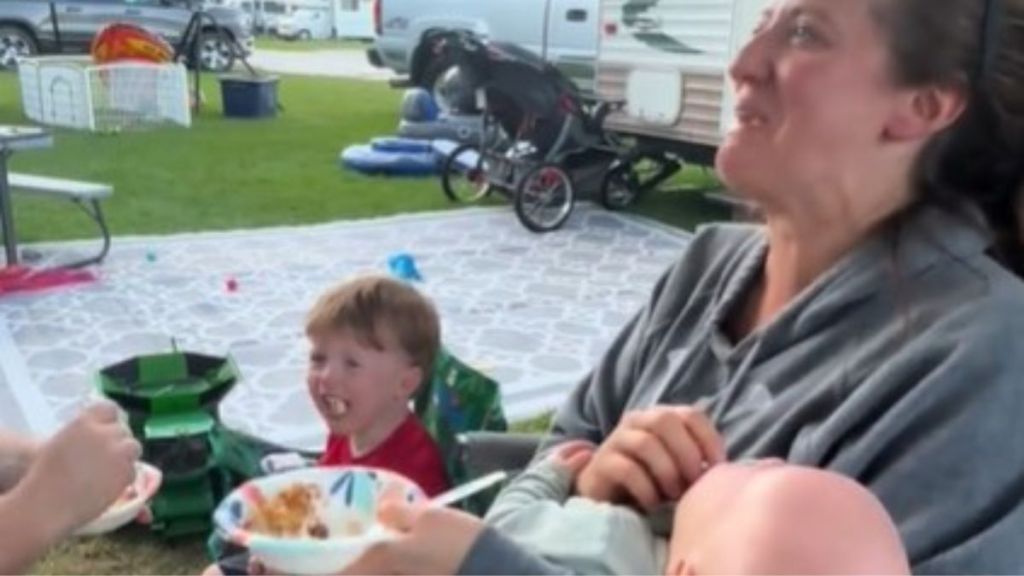 Image shows a family at a campground with a mom rocking holding one screaming child while a toddler has a tantrum next to her.