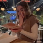 A woman sits at a table at a restaurant. She's reading a card that she holds with one hand. With her other hand she covers her mouth. She looks emotional