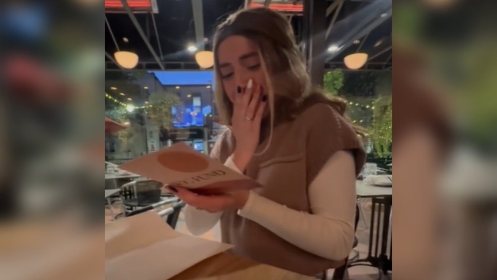 A woman sits at a table at a restaurant. She's reading a card that she holds with one hand. With her other hand she covers her mouth. She looks emotional