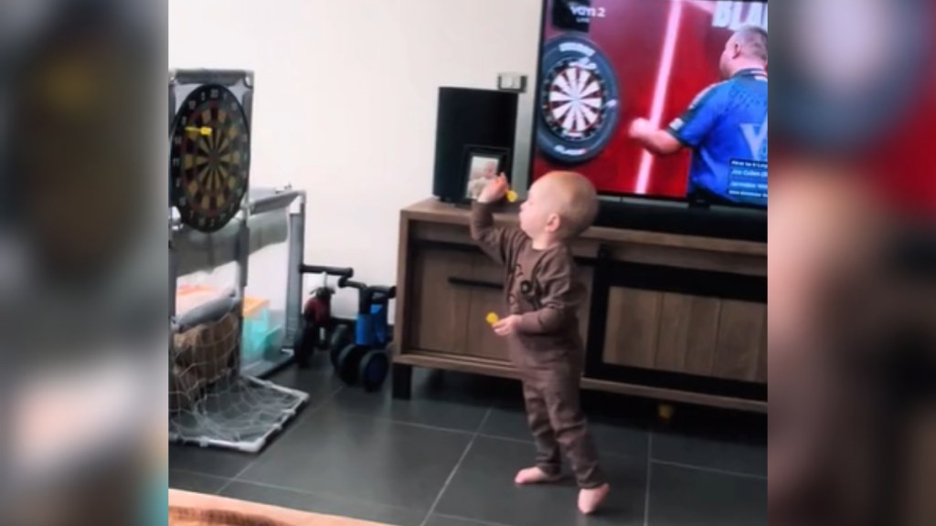 A baby stands near a TV that is showing a professional game of darts. The baby looks away from the TV and toward a real dart board in his living room. He poses as he prepares to throw the magnetic dart.