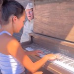 Side view of a young woman playing a piano that's outdoors. A man sits next to the piano, smiling as he watches her play