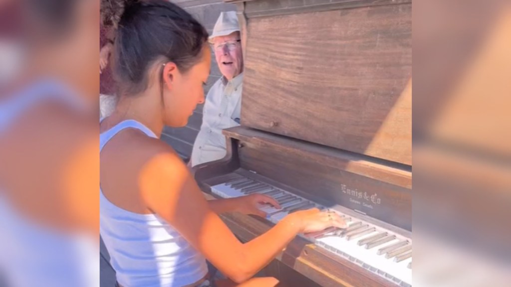 Side view of a young woman playing a piano that's outdoors. A man sits next to the piano, smiling as he watches her play