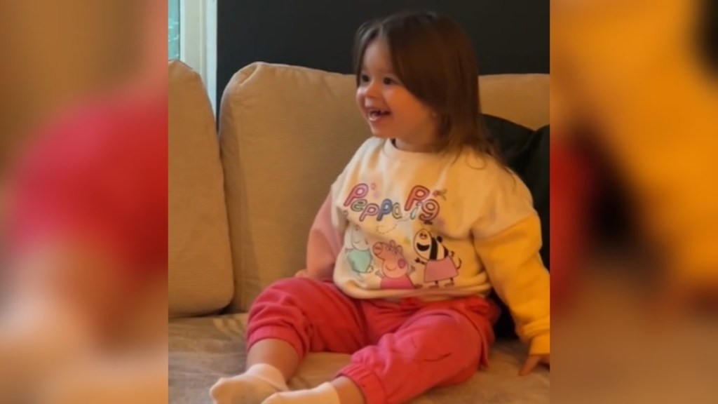 Close up of a toddler sitting on a couch. She smiles, eyes wide with happiness, as she looks over at something that can't be seen in this photo