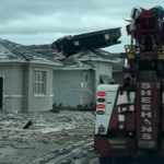 View from behind of a tow truck driving past a destroyed house with a dumpster on the roof