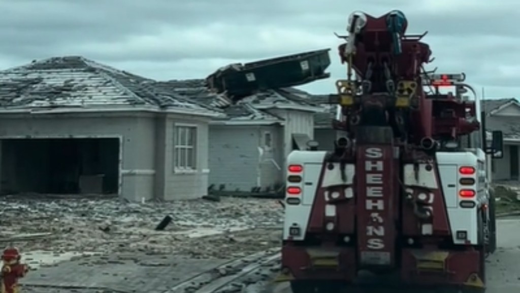 View from behind of a tow truck driving past a destroyed house with a dumpster on the roof