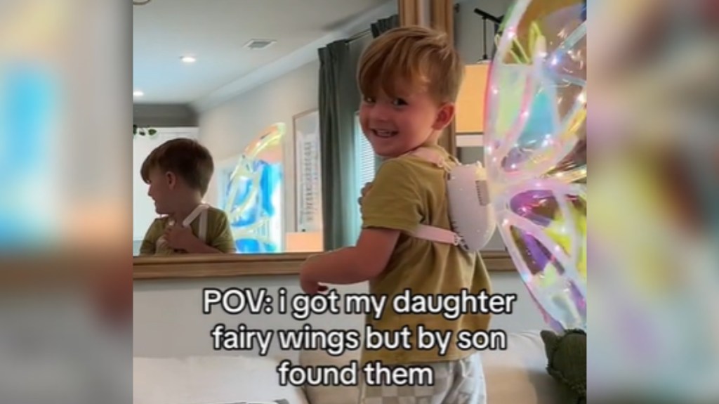 A little boy stands on a couch to look at the mirror on the wall. He's wearing iridescent fairy wings with lights. He looks back at the camera and gives a sweet smile. Text on the image reads: POV: I got my daughter fairy wings but my son found them