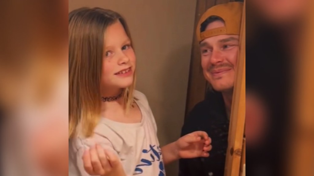 View of a little girl and Dad looking into a mirror. While the girl smiles, admiring her hair, Dad looks at her, tears in his eyes as he gives an emotional smile.