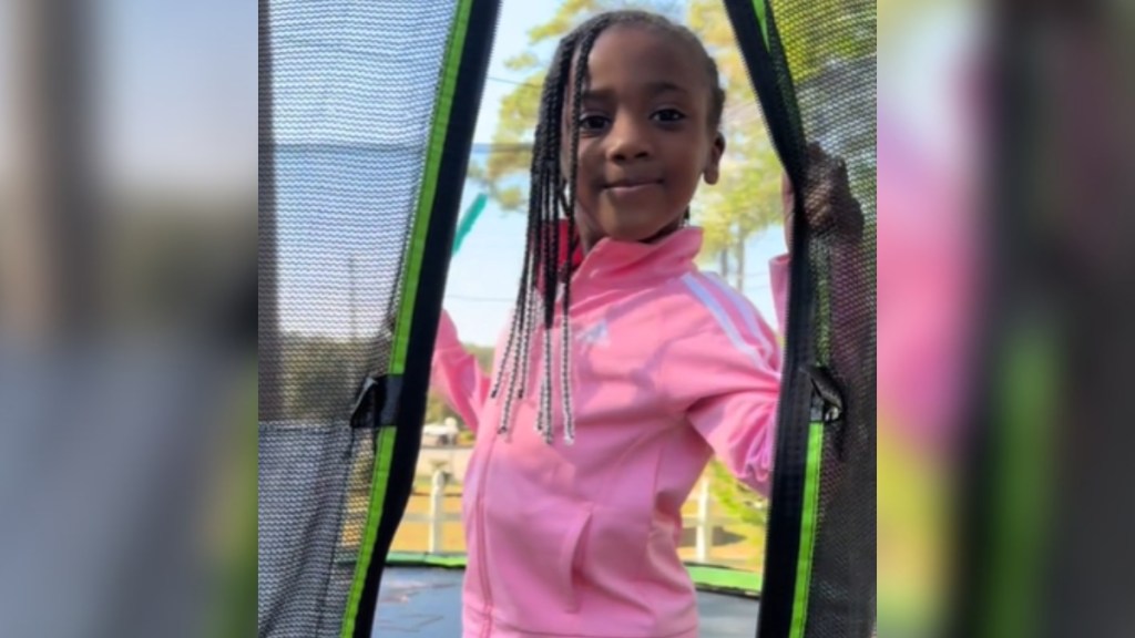 A little girl gives a close-lip smile, looking proud. She's standing on a trampoline and is holding back the mesh netting