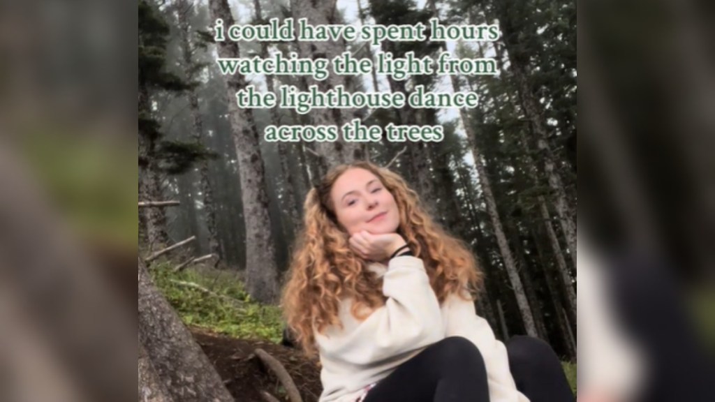 A woman smiles as she poses with her head rested on one of her hands. She sits in a forest. Text on the image reads: I could have spent hours watching the light from the lighthouse dance across the trees