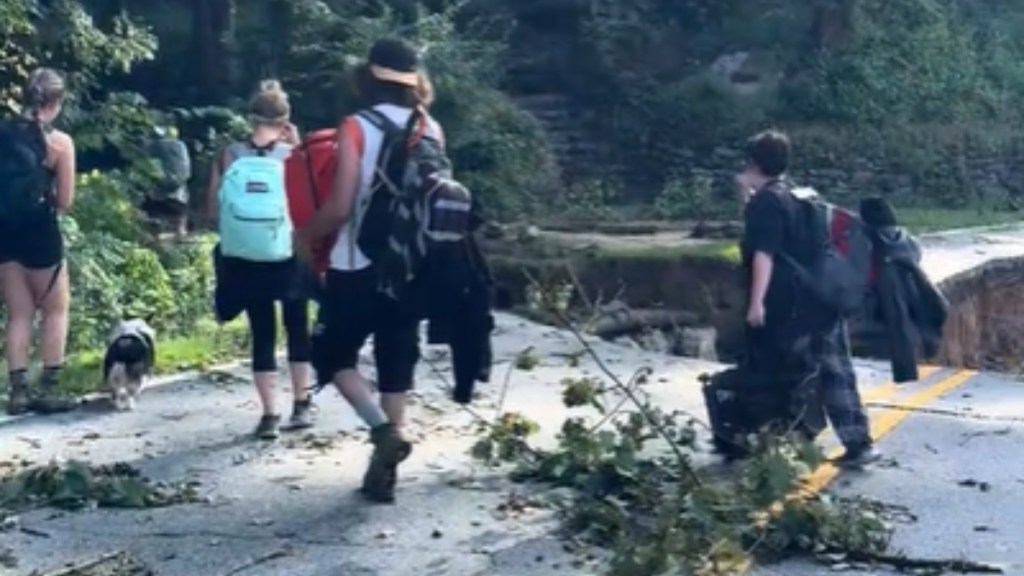 A group of people wearing backpacks walks along a road with debris and nearby a massively destroyed part of the road that leads to water