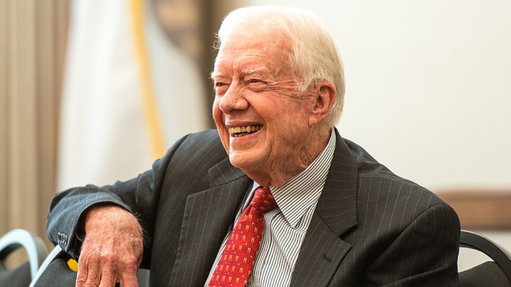 Jimmy Carter smiles wide as he sits in a chair, one arm resting on the chair next to him