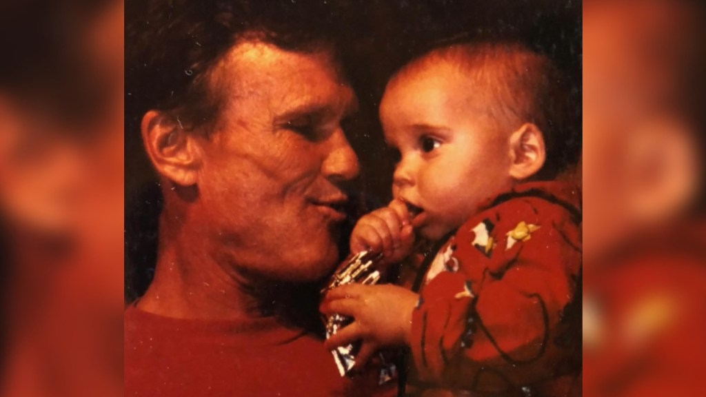 Old photo of a young Kris Kristofferson smiling as he holds a baby in his arms. The baby is Willie Nelson's son, Lukas Nelson.