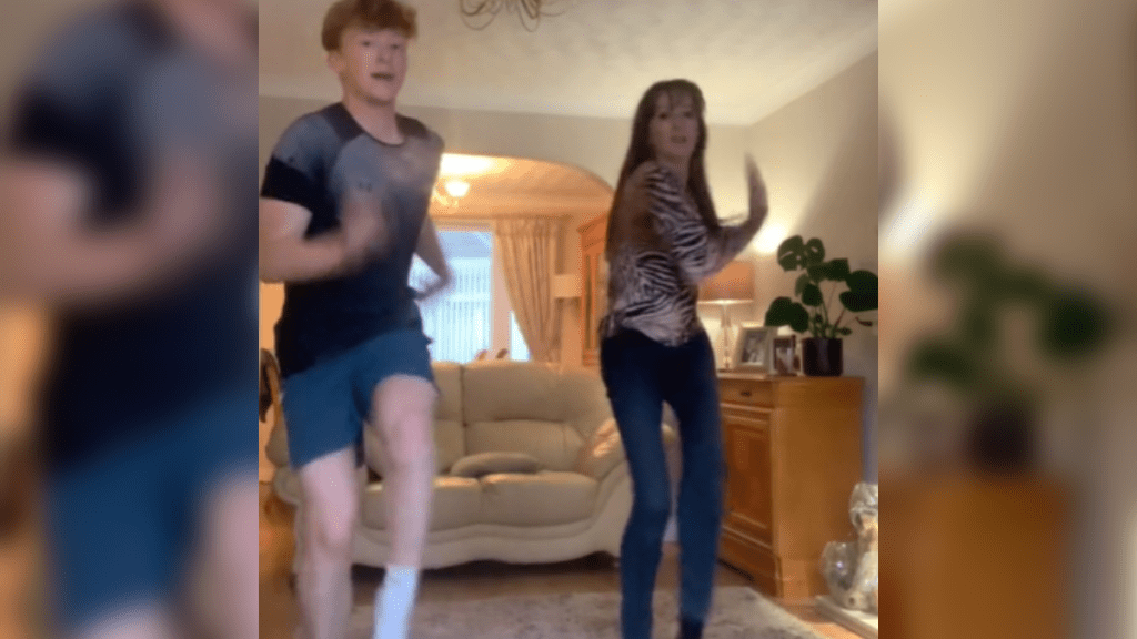 A young man and his mom are mid-dance and doing the same move. Both of them look into the camera.