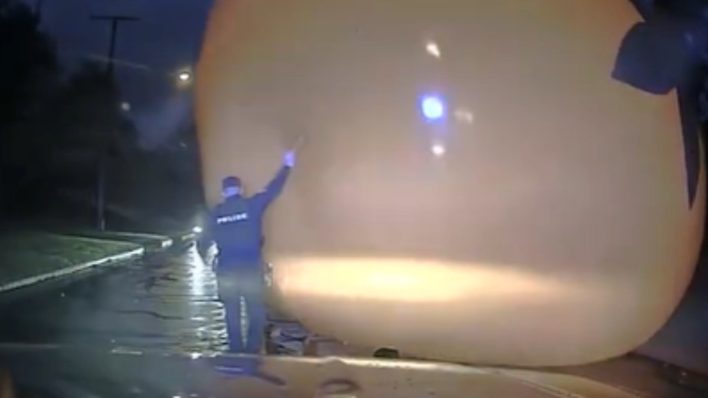 Dashcam footage of a police officer approaching a massive Halloween pumpkin decoration. The officer holds up an arm to stop the decor from running into him