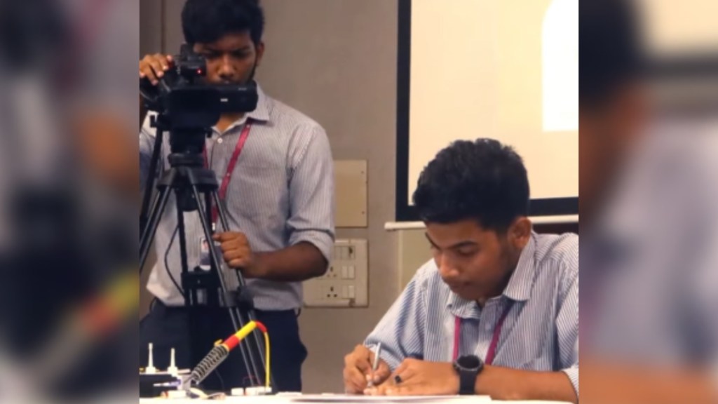 A man concentrates while he does something at a table. Another man stands next to him with a large camera, recording the moment