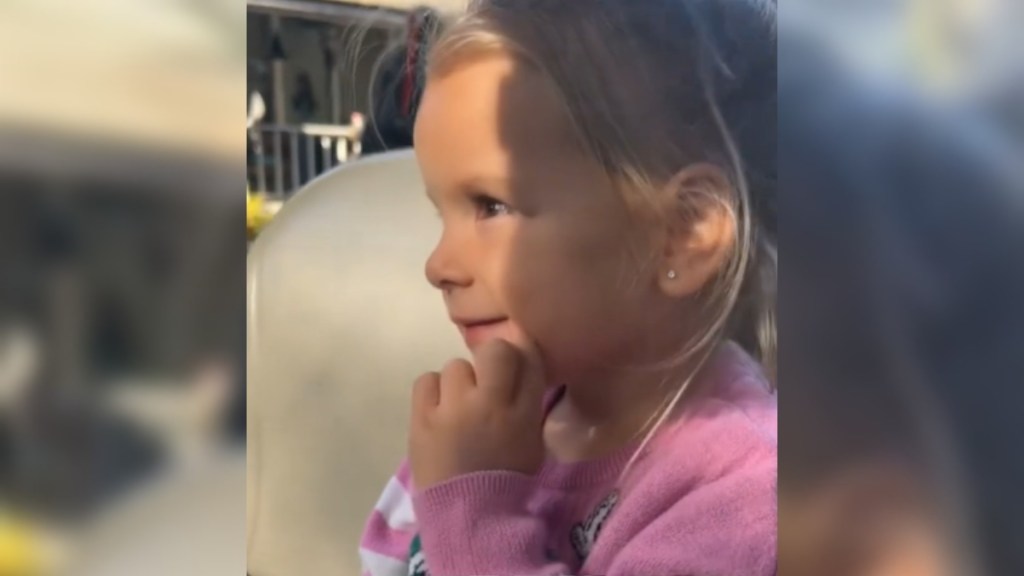 Close up of a little girl sitting in a partially shaded area outside. She holds a hand to her chin as she smiles, entranced by something not shown in the photo.