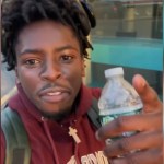 Close up of a man talking to the camera. He's holding a water bottle while also pointing to the camera. He's walking outside