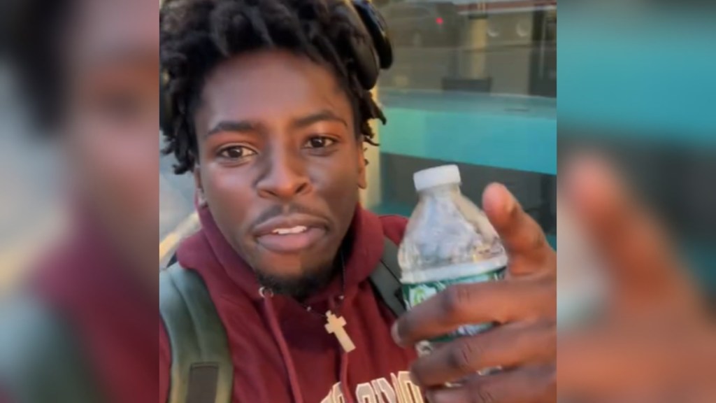 Close up of a man talking to the camera. He's holding a water bottle while also pointing to the camera. He's walking outside