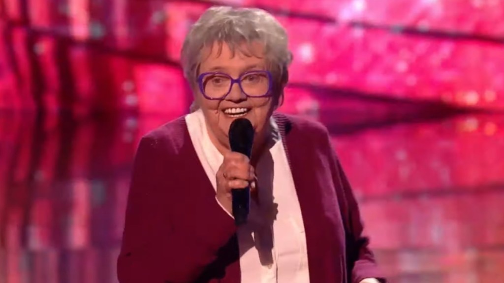 An elderly woman smiles wide as she sings into a mic on "The Voice UK"