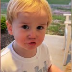 Close up of a toddler making an adorable face as he talks. He's standing on a front porch