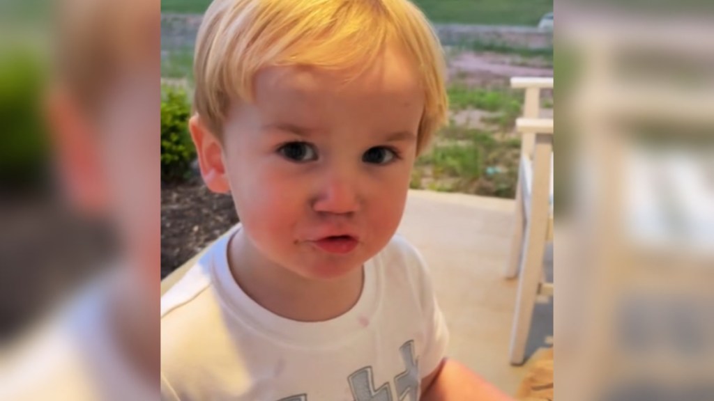 Close up of a toddler making an adorable face as he talks. He's standing on a front porch