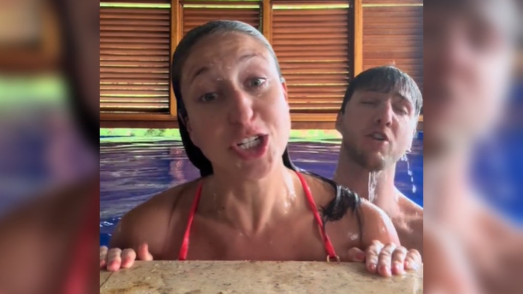 Close up from the side of a pool. A woman rests her hands on the edge of the pool, singing into the camera. A man stands in the pool just behind her, also singing, but his are closed