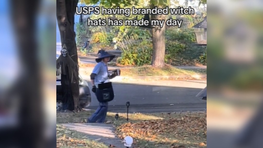 From a distance, a USPS driver walks along a sidewalk while wearing a witch hat. Text on the image reads: USPS having branded witch hats has made my day