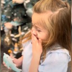 Close up of a little girl standing in front of a Christmas tree. While she holds an ornament in one hand, she uses her other hand to cover her mouth. Her eyes are a bit shut, and she looks emotional