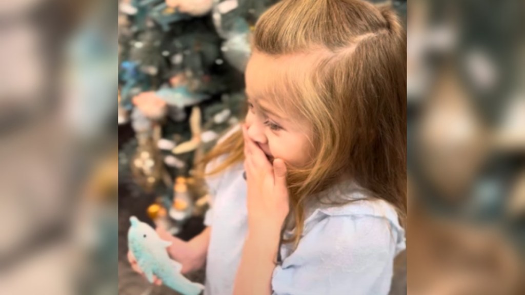 Close up of a little girl standing in front of a Christmas tree. While she holds an ornament in one hand, she uses her other hand to cover her mouth. Her eyes are a bit shut, and she looks emotional