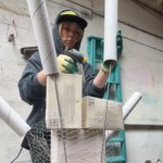 A woman wearing gloves adds to a display made up of boxes and tubes. She holds one tube with one hand and, with the other, a hot glue gun.