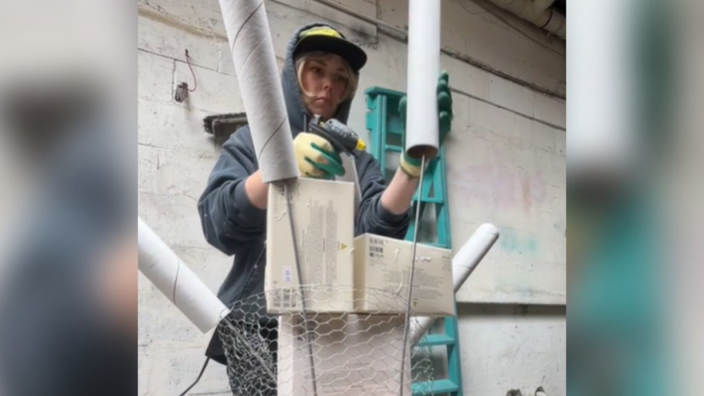 A woman wearing gloves adds to a display made up of boxes and tubes. She holds one tube with one hand and, with the other, a hot glue gun.