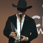 Cody Johnson speaks into a mic as he holds his CMA award for Album of the Year. He wears a hat that partially obscures his face in a shadow