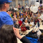 A man leads a crowd of elementary students in a song. A pit bull in a birthday hat sits in a cart, happily taking in the love