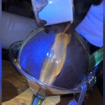 Close up of a dessert. A late chocolate ball sits in a large drinking glass. Warm liquid is being poured onto the chocolate.