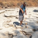 A man stands in a dried up riverbed, using a long pole to point out dinosaur tracks