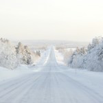 View of a snow-covered road. Snow-covered trees line the road. The sky in the distance is covered in a bright hazy white/grey