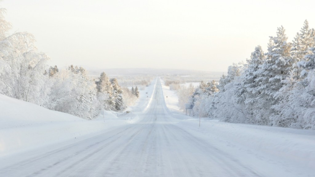 View of a snow-covered road. Snow-covered trees line the road. The sky in the distance is covered in a bright hazy white/grey