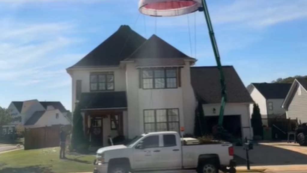 View from the street of a massive two-story home. A machine is being used to lift something onto the home, but the object can't be fully seen in this image. A person supervises from the ground.