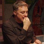 Alan Tudyk sits at a table and looks down as he talks, one hand held close to his mouth. He has a serious expression on his face