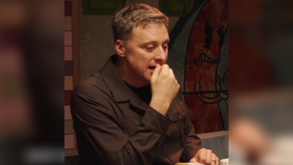 Alan Tudyk sits at a table and looks down as he talks, one hand held close to his mouth. He has a serious expression on his face