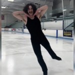 An ice skater poses with his hands under his face as he smiles. One foot is raised in the air, slightly behind him, as he skates