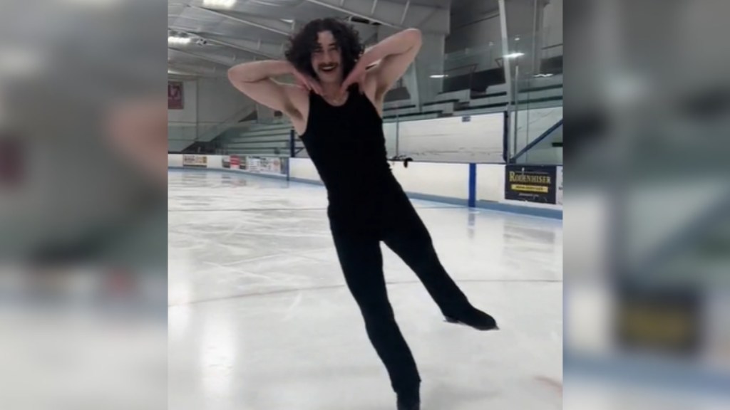 An ice skater poses with his hands under his face as he smiles. One foot is raised in the air, slightly behind him, as he skates