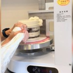 Close up of an "icing machine." A cake is on the machine and icing is being placed on it while it spins. Someone's hands can be seen squeezing the icing on.