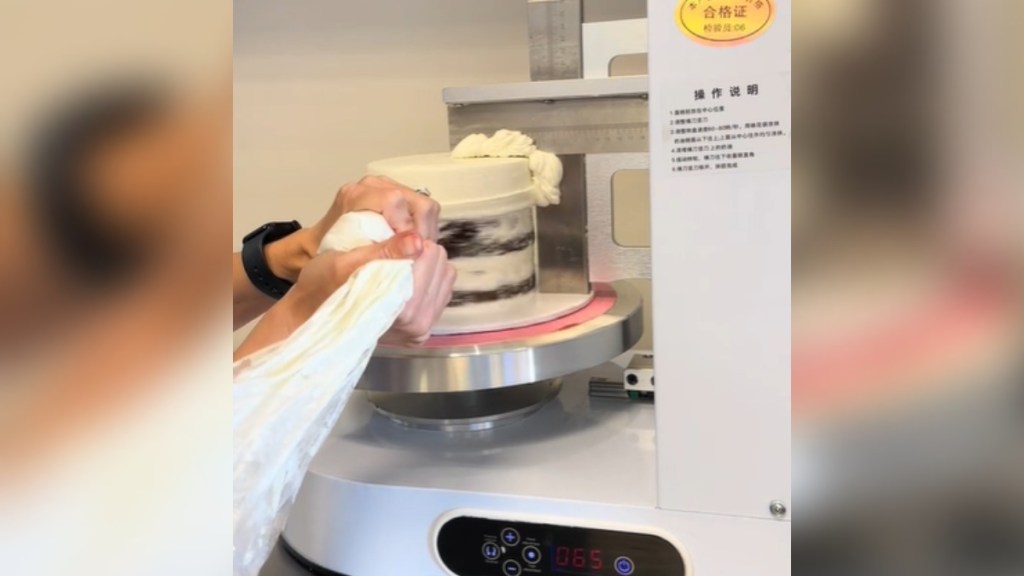 Close up of an "icing machine." A cake is on the machine and icing is being placed on it while it spins. Someone's hands can be seen squeezing the icing on.