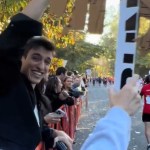 View of bystanders cheering on runners in the 2024 NYC Marathon. The person holding the camera uses their one free hand to hold up a sign we can't see from this angle. A friend next to her does the same. He smiles wide, laughing, as he looks back at her. The runners run past nearby