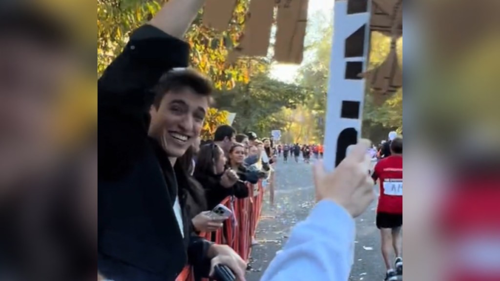 View of bystanders cheering on runners in the 2024 NYC Marathon. The person holding the camera uses their one free hand to hold up a sign we can't see from this angle. A friend next to her does the same. He smiles wide, laughing, as he looks back at her. The runners run past nearby