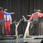 Four treadmills are set up on a stage. Four teens stand on one each. The two closes to the camera have their backs facing us. The two teens behind them face us. They each have their arms stretched out in a cool formation